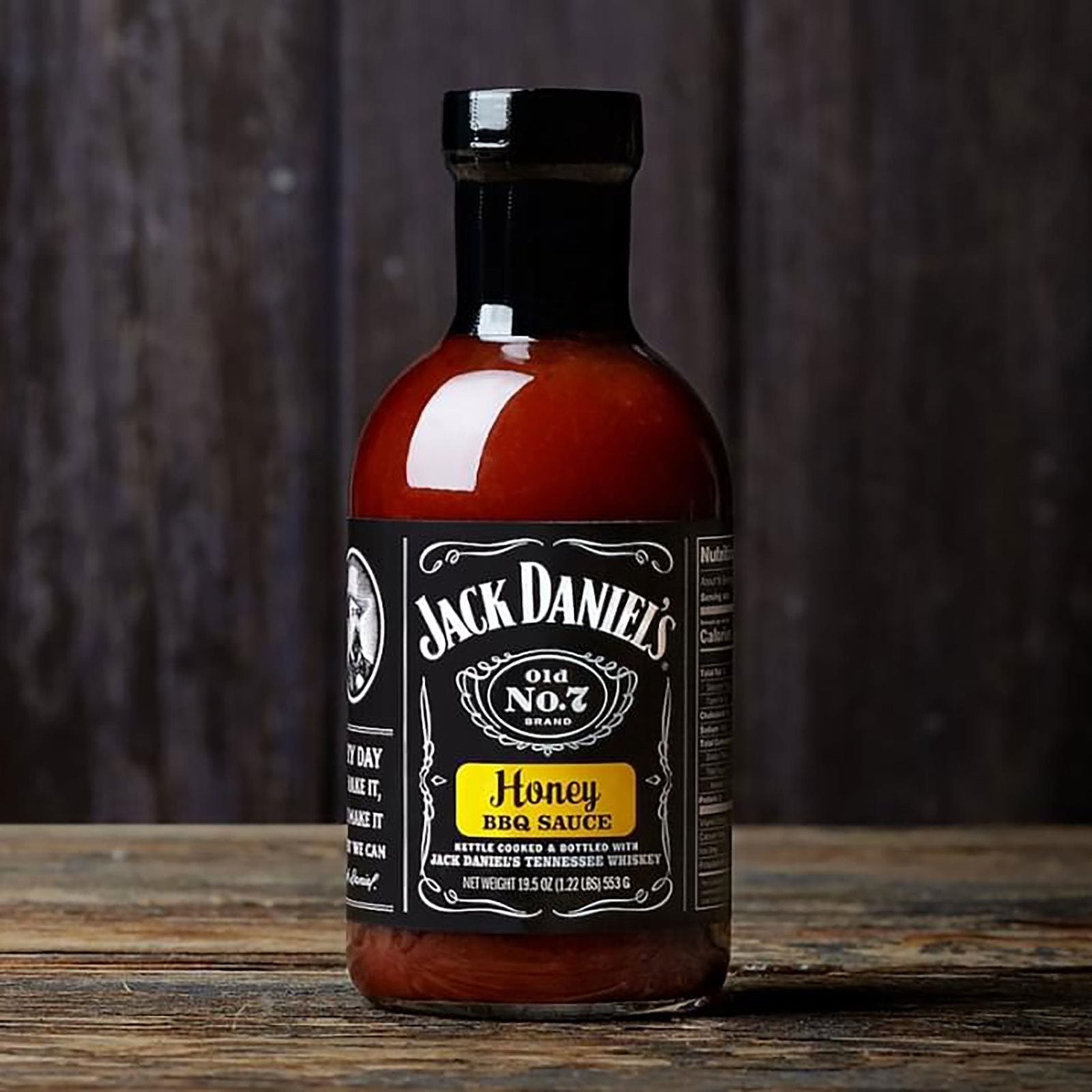 En 473 ml flaske Jack Daniel's BBQ Honey hviler på en treflate med en mørk trebakgrunn som fremhever den fyldige honningsmaken inspirert av Jack Daniel's Tennessee-whiskey.