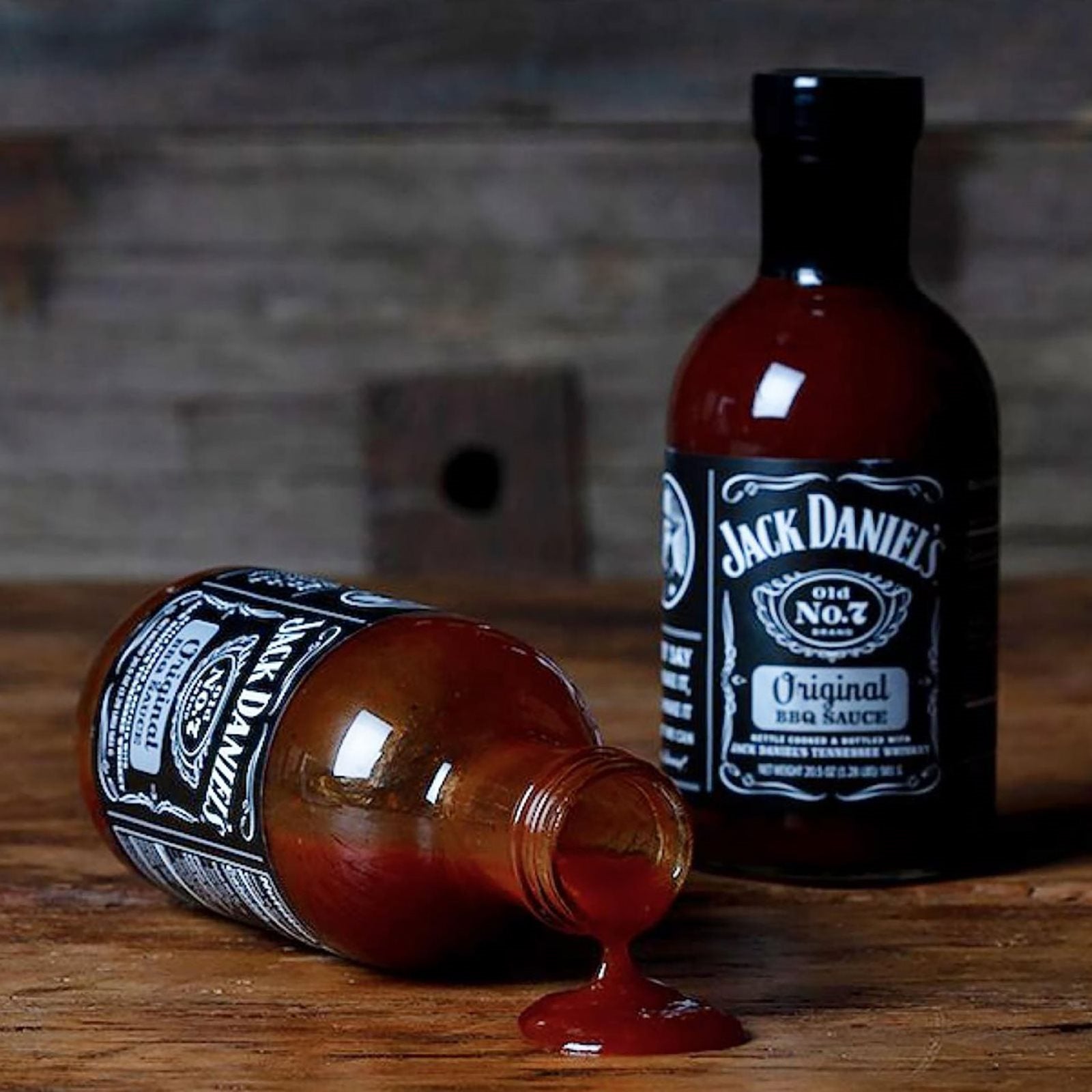 To flasker Jack Daniel's BBQ Original (473 ml) på en treflate - den ene står oppreist, den andre er veltet med den karakteristiske Tennessee Whiskey-sausen som renner ut.