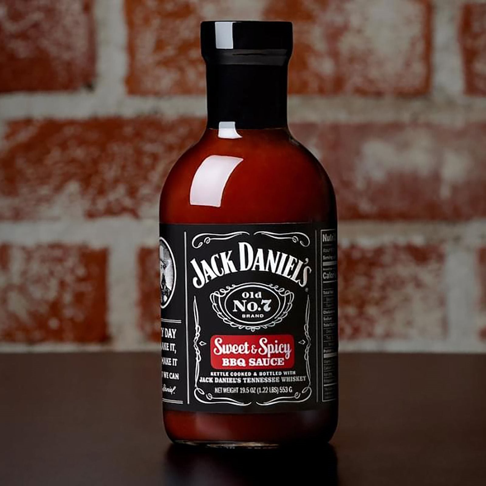 En 473 ml flaske Jack Daniel's BBQ Sweet & Spicy saus står på en mørk flate foran en murvegg.