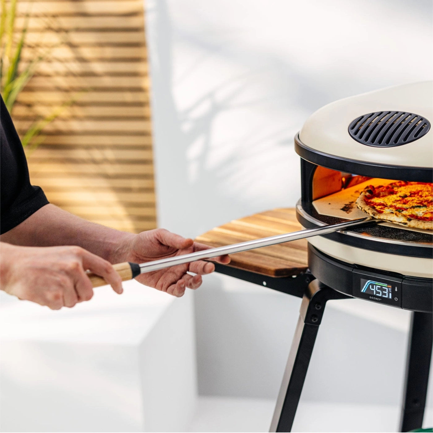 En person bruker en «Gozney Balance Turning Peel» til å ta ut pizza fra en utendørsovn med digital temperaturvisning.