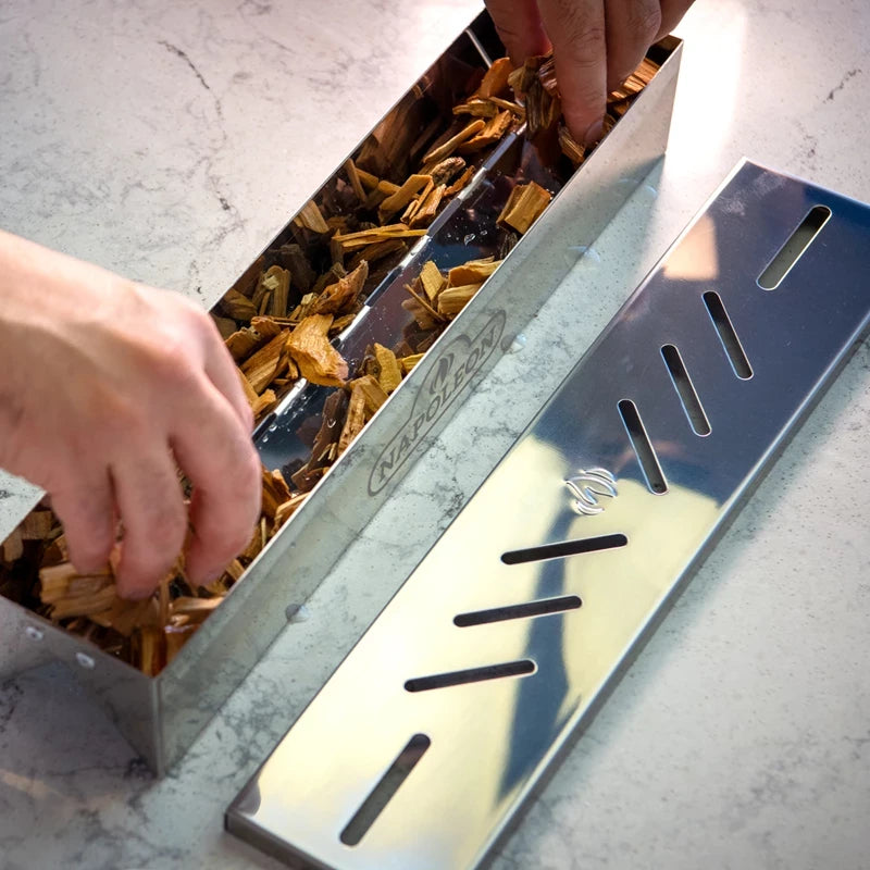 Hendene legger flis i en Napoleon Sear Plates Smoker Box - røykeeske i rustfritt stål med slisset lokk på en lys benkeplate, perfekt for å legge røyksmak til gassgrill.