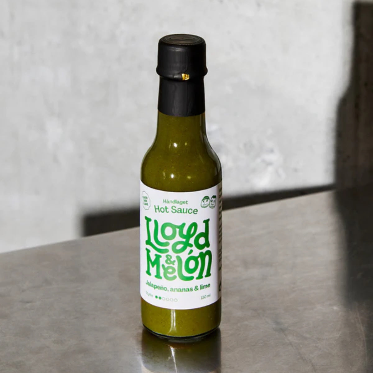 En flaske Jalapeño & Ananas Hot Sauce | Lloyd & Melón hviler på en grå flate mot en ensfarget bakgrunn.