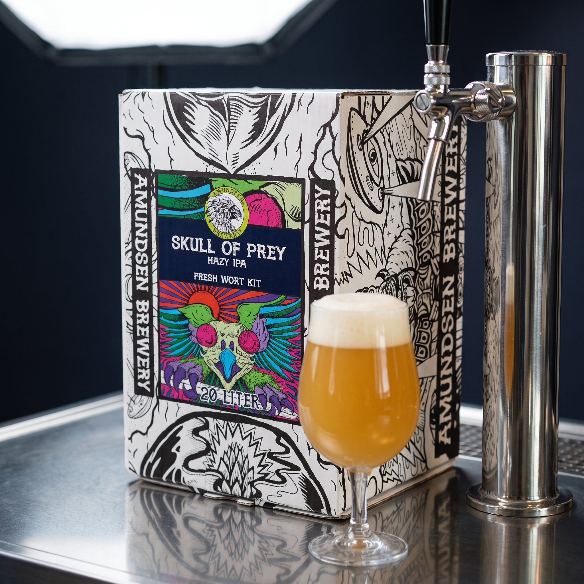 En eske med påskriften «Amundsen Skull of Prey Hazy IPA – 20L Fresh Wort Kit» står ved siden av en ølkran og et glass med hazy IPA på en metallflate.