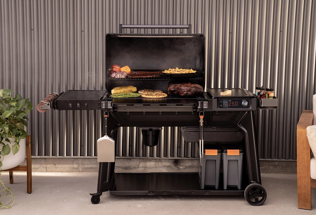 En stor Traeger Woodridge Pro (Forhåndsbestilling) pelletsgrill med åpent lokk viser sydende grønnsaker, mais og kjøtt på ristene. Grillverktøy og oppbevaringsrom er plassert under, og den smarte WiFIRE-teknologien sørger for enkel kontroll.