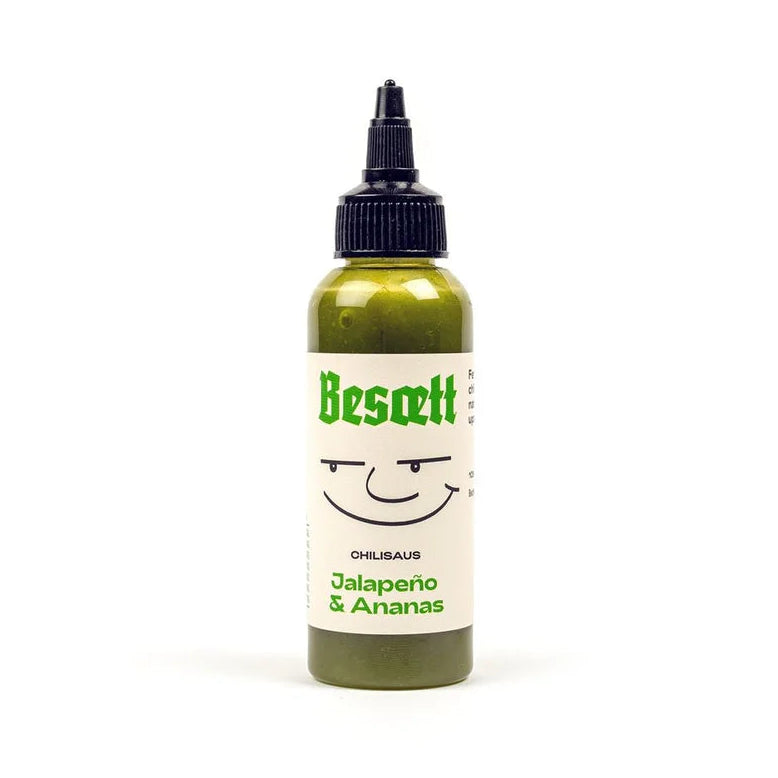 En liten flaske med «Besætt Jalapeño & Ananas Fermentert Chilisaus», med en grønn etikett med et enkelt smilende ansikt og en svart påfyllingshette, mot en hvit bakgrunn.