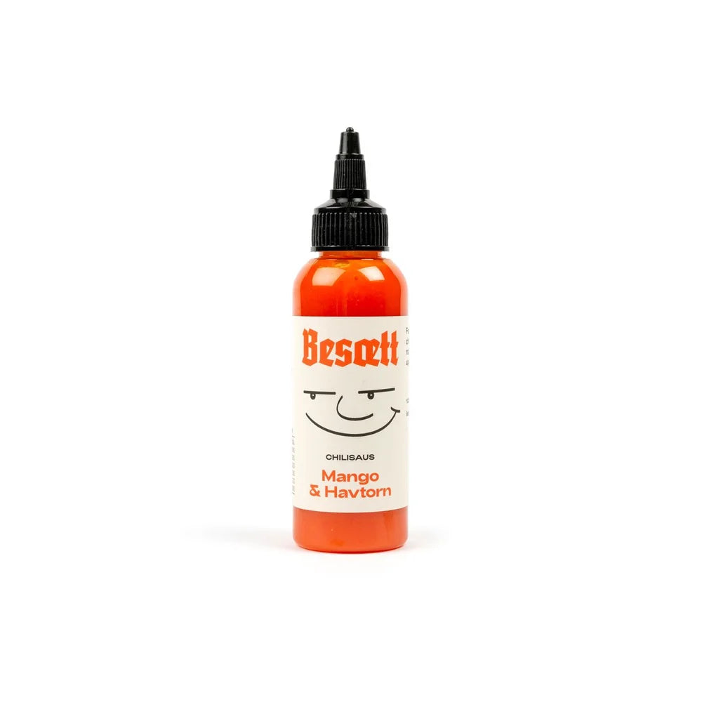 En flaske «Besætt Mango & Havtorn Fermentert Chilisaus», med en illustrasjon av et smilende ansikt på forsiden og en Carolina Reaper-chili, isolert på hvit bakgrunn.