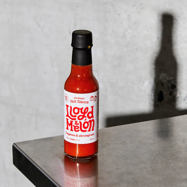 En flaske Cayenne & Sitrongress Hot Sauce fra Lloyd & Melón hviler på en metallflate med en betongvegg i bakgrunnen.
