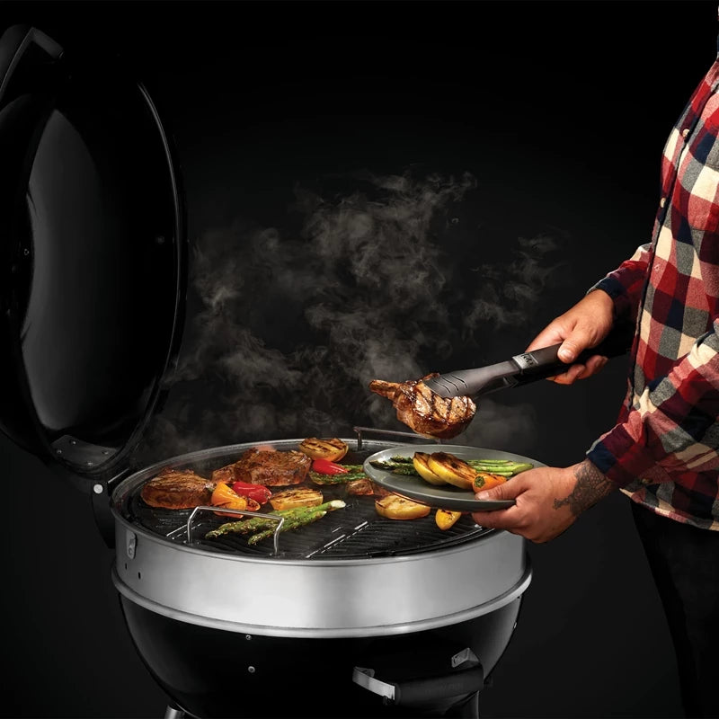 Ved grilling av grønnsaker og kjøtt på en Napoleon PRO22 Kulegrill 57 cm holder personen i en tang og en tallerken mens dampen stiger deilig opp fra maten.