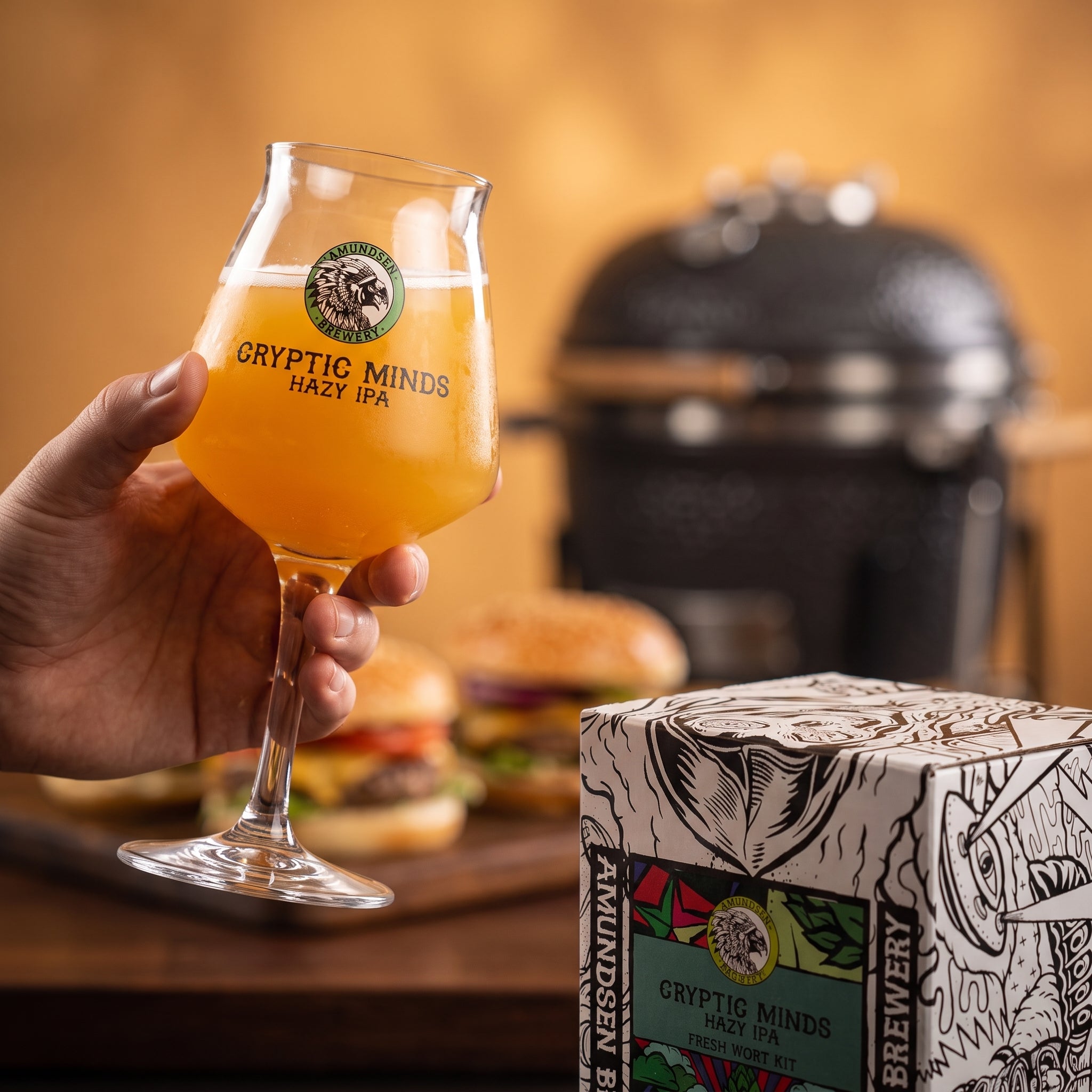 En hånd holder et glass Amundsen Cryptic Minds Hazy IPA; bak stekes det burgere på grillen, mens en eske med Amundsen Cryptic Minds Hazy IPA – 20L Fresh Wort Kit står tydelig frem i forgrunnen.