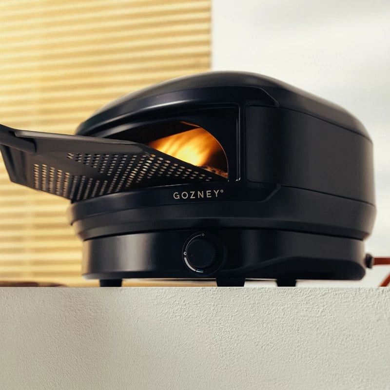 The Gozney Arc Lite, Sort Pizzaovn vises med en pizzaspade delvist indsat og flammer synlige inde i denne kompakte ovn.