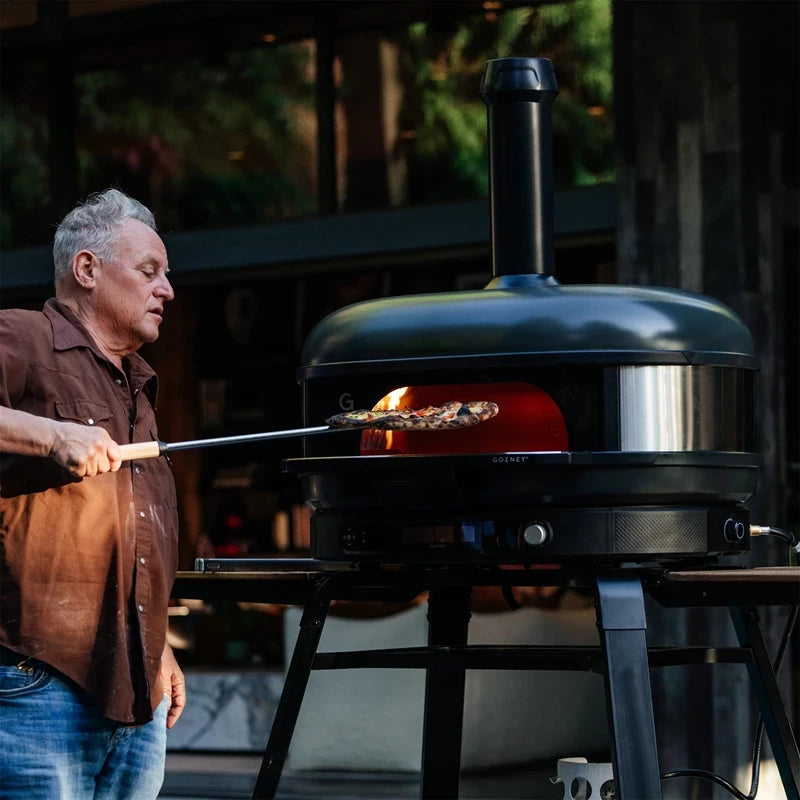 En person bruker et verktøy med langt skaft til å plassere eller fjerne pizza fra en Gozney Dome Gen 2 XL - Off Black - Multi Fuel Pizzaovn, som er plassert på et stativ på et utekjøkken.