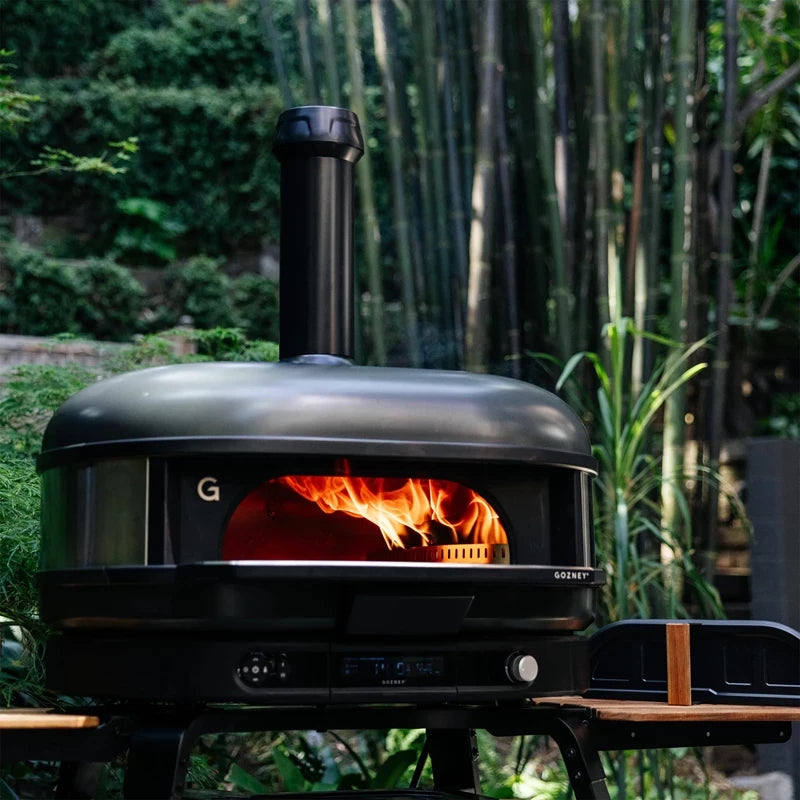 En Gozney Dome Gen 2 XL - Off Black - Multi Fuel Pizzaovn med skorstein og synlige flammer inni er satt opp i et frodig utekjøkken i en hage omgitt av bambus og grønne planter.