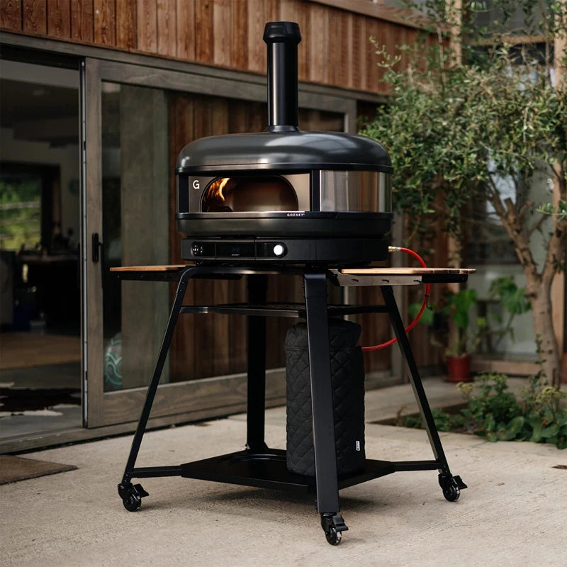 Gozney Dome Gen 2 XL - Off Black - Multi Fuel Pizzaovn med skorstein står på en metallvogn på hjul med åpne sidebord og en propantank under, plassert på en uteplass ved huset - ideelt for ethvert utendørs kjøkken.