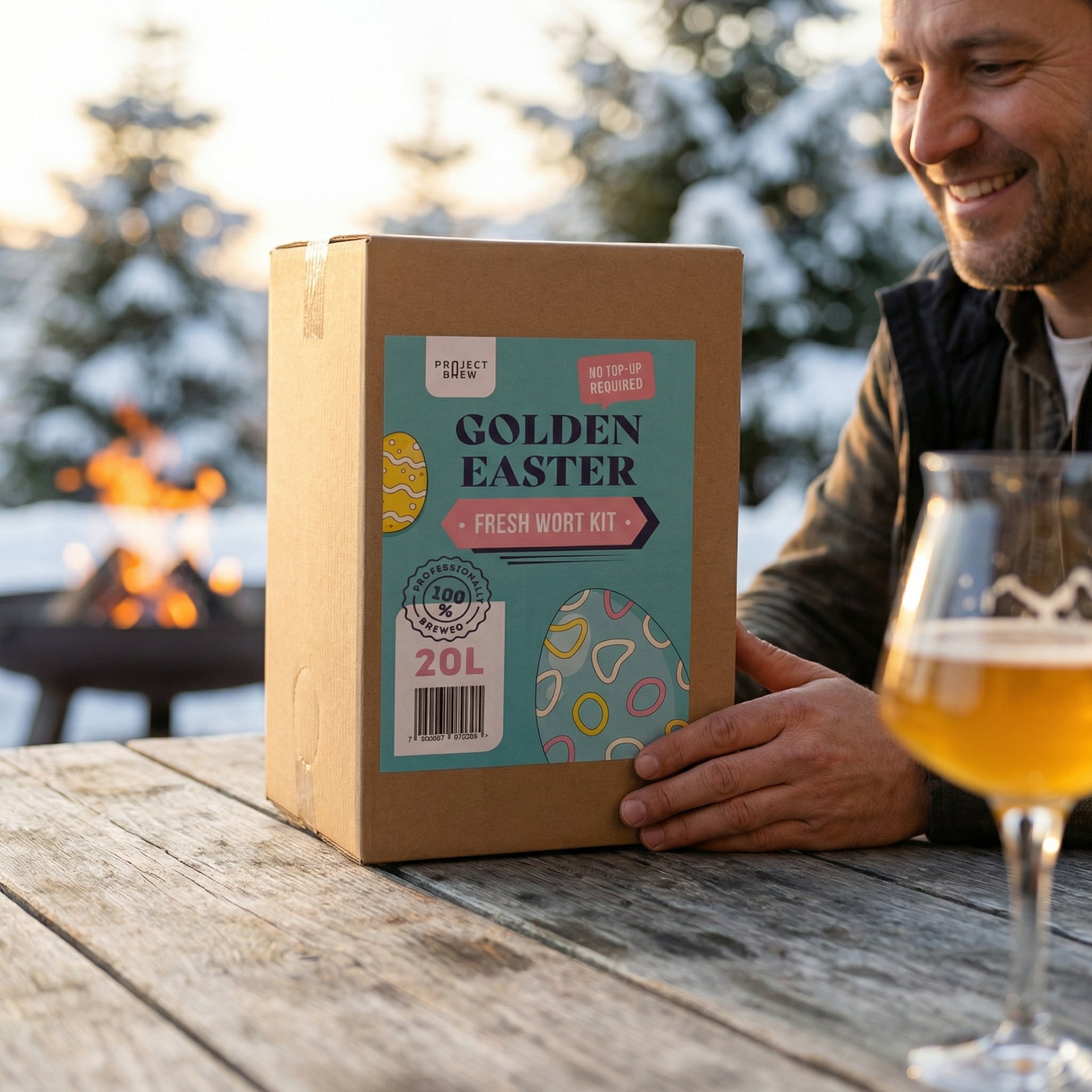 En mann holder en Golden Easter - 20L Fresh Wort Kit-boks ved et utendørsbord med et glass belgisk witbier; en bålplass og snødekte trær skaper et koselig bakteppe, perfekt for å nyte dette påskeølet (også godt som sommerbrygg).