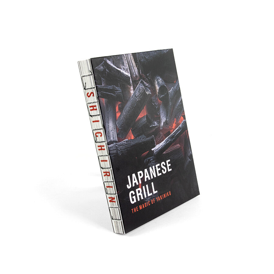 Cookbook Japanese Grill YAKINIKU, Shichirin Kokebok, står oppreist på en hvit bakgrunn med et brennende trekulldeksel - ideelt for alle som ønsker å mestre autentiske japanske grillteknikker.