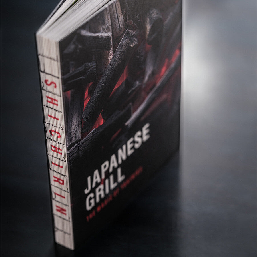 Cookbook Japanese Grill YAKINIKU, Shichirin Kokebok står oppreist på en mørk overflate, med et deksel som viser forkullet treverk og glør som fanger essensen av det autentiske japanske grillkjøkkenet.