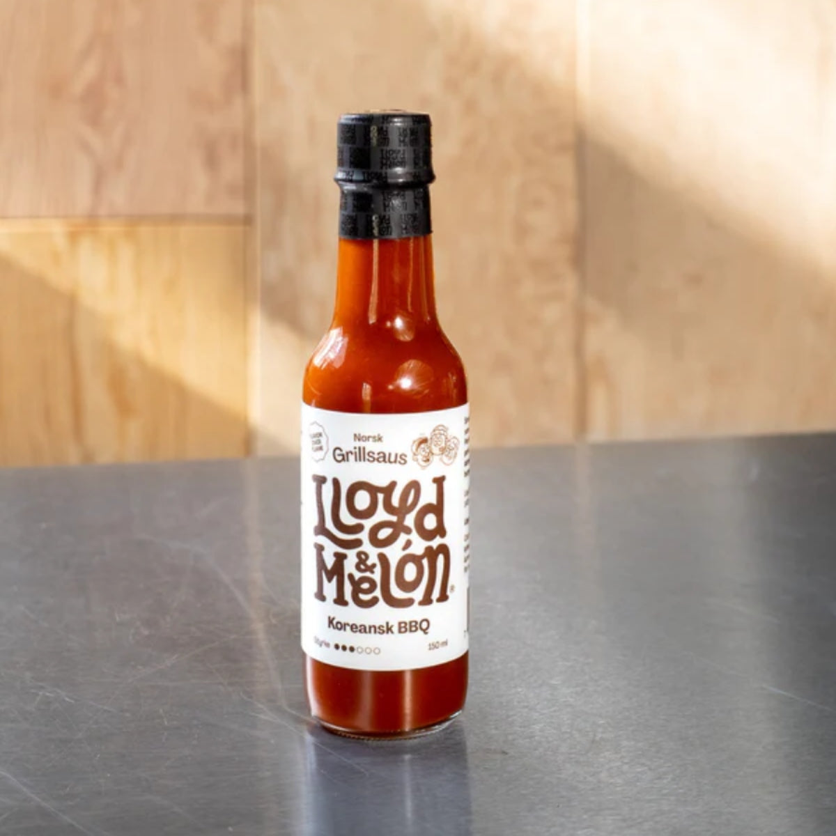 En flaske Koreansk BBQ Hot Sauce fra Lloyd & Melón, med gochujang og glutenfri soyasaus, er plassert på en grå benkeplate med en lys trebakgrunn.