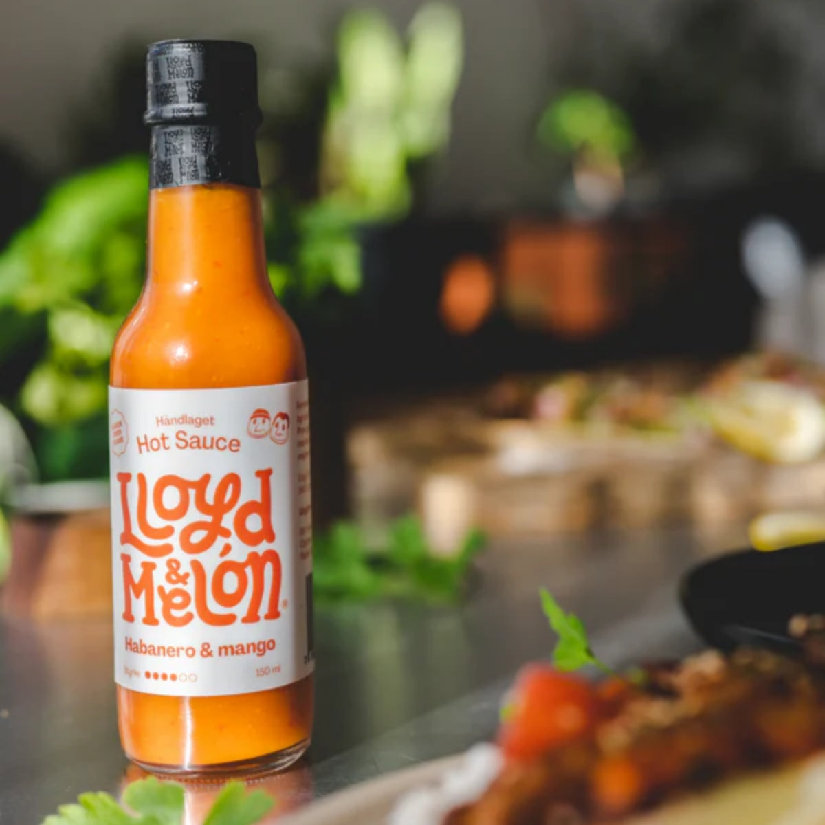 En flaske Habanero & Mango Hot Sauce | Lloyd & Melón står på et bord, omgitt av mat og uskarpt grønt, og fremhever en av de håndlagde hot sausene deres.