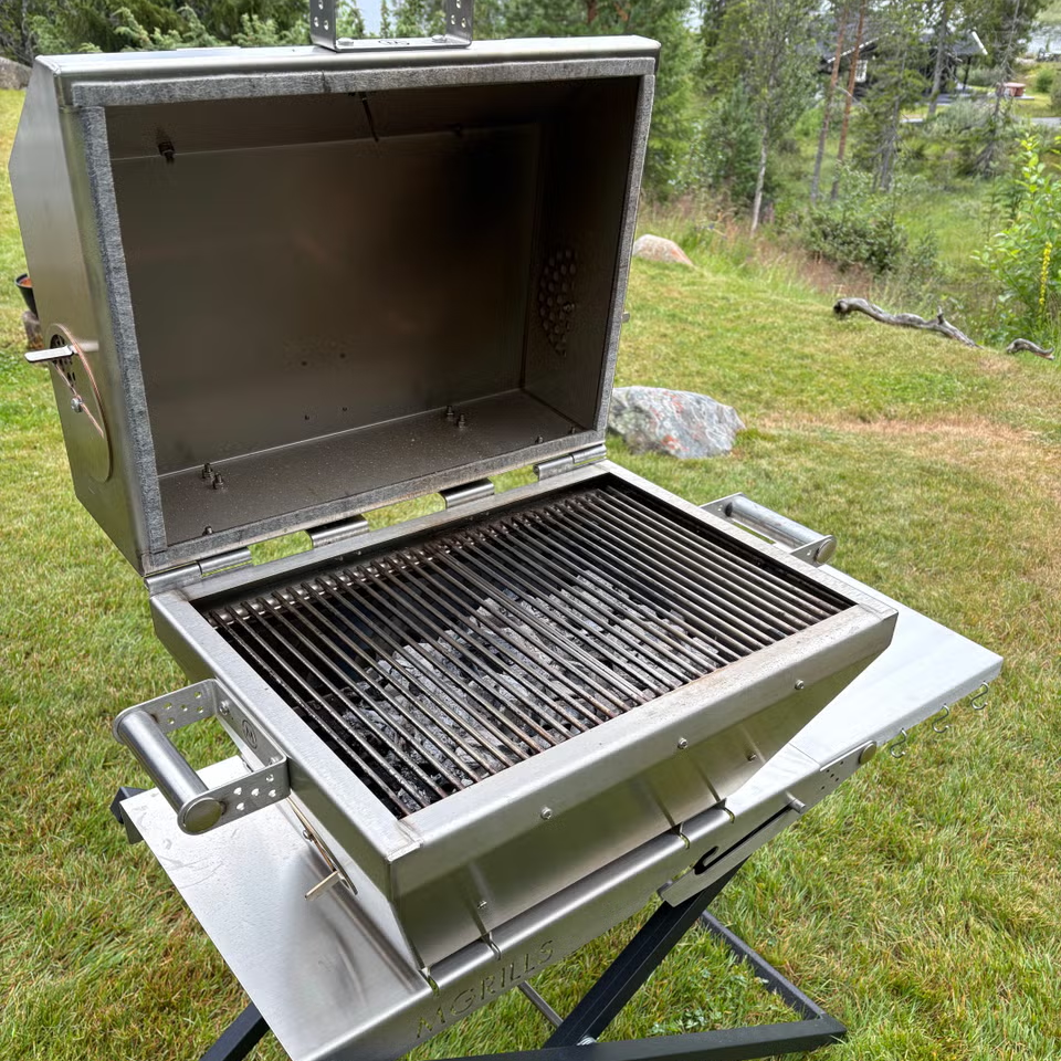 En M16 Cookoff Grill i rustfritt stål – M-Grills – Kullgrill med temperaturkontroll står på en plen med lokket åpent, og viser rene grillrister samt trær og steiner bak den.