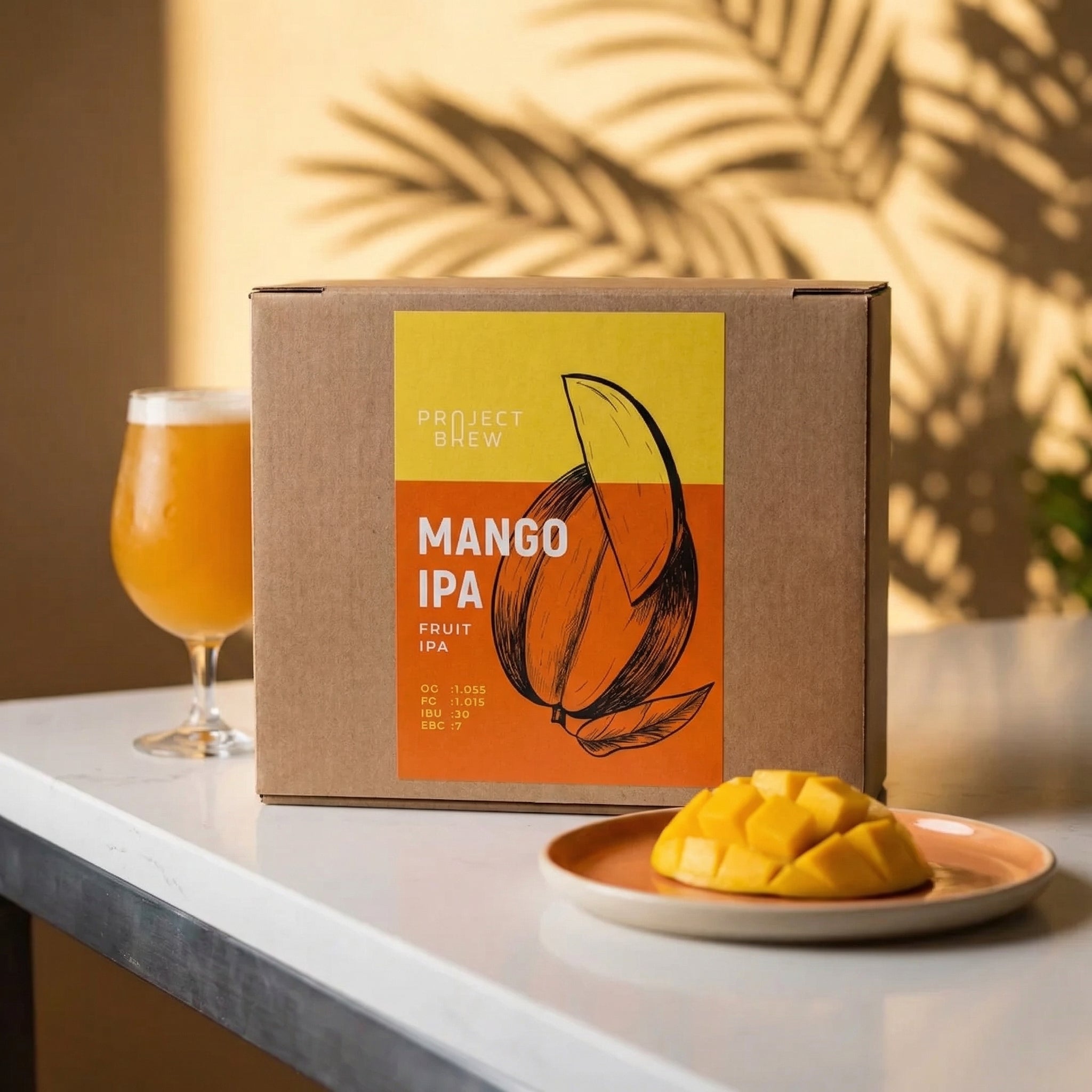 En eske merket Mango IPA - allgrain ølsett står på et hvitt bord ved siden av et glass fruktig IPA og en tallerken med mangoskiver, med sollys og planteskygger i bakgrunnen.