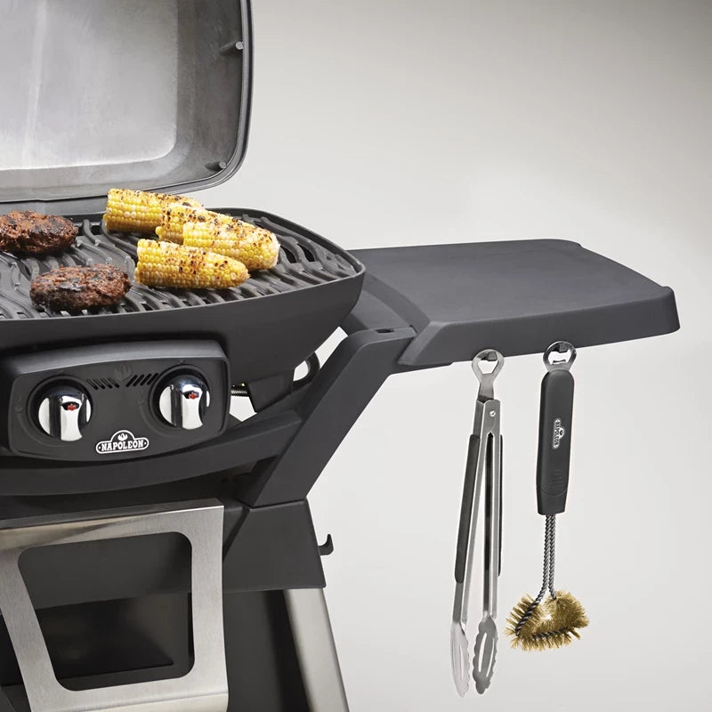 En gassgrill med mais og burgere som steker på risten; et Napoleon Redskapsett med grilltang, spade og børste henger fra kroker på sidehyllen.