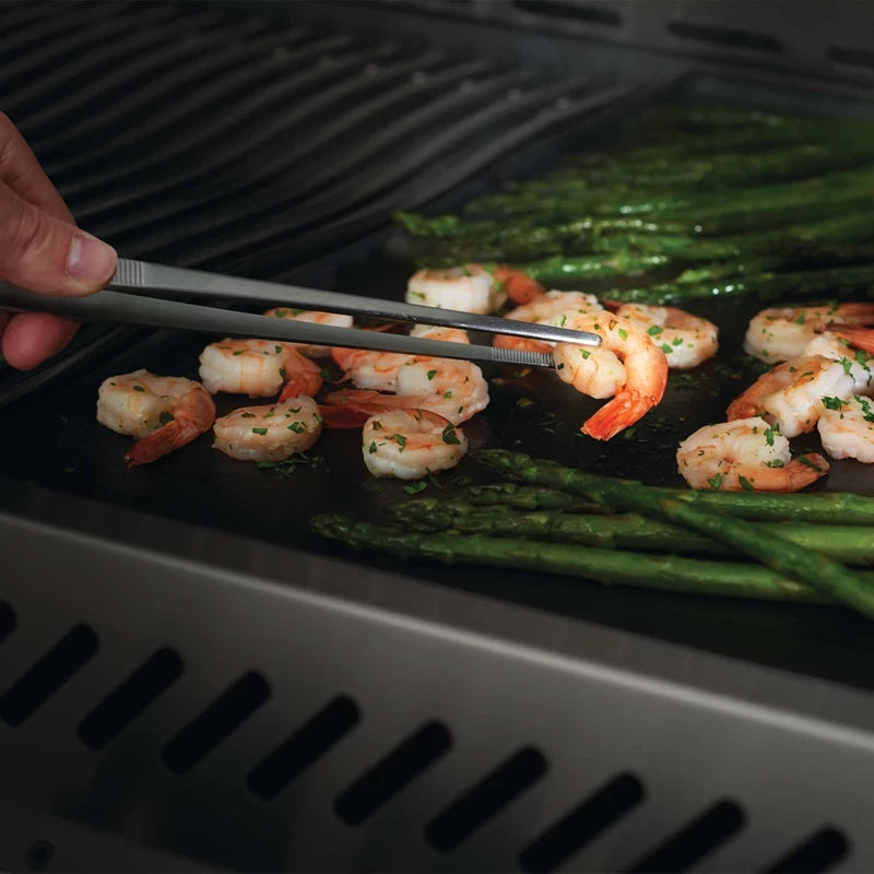 En hånd bruker Napoleon matpinsett i rustfritt stål til å grille reker og asparges på en grill.