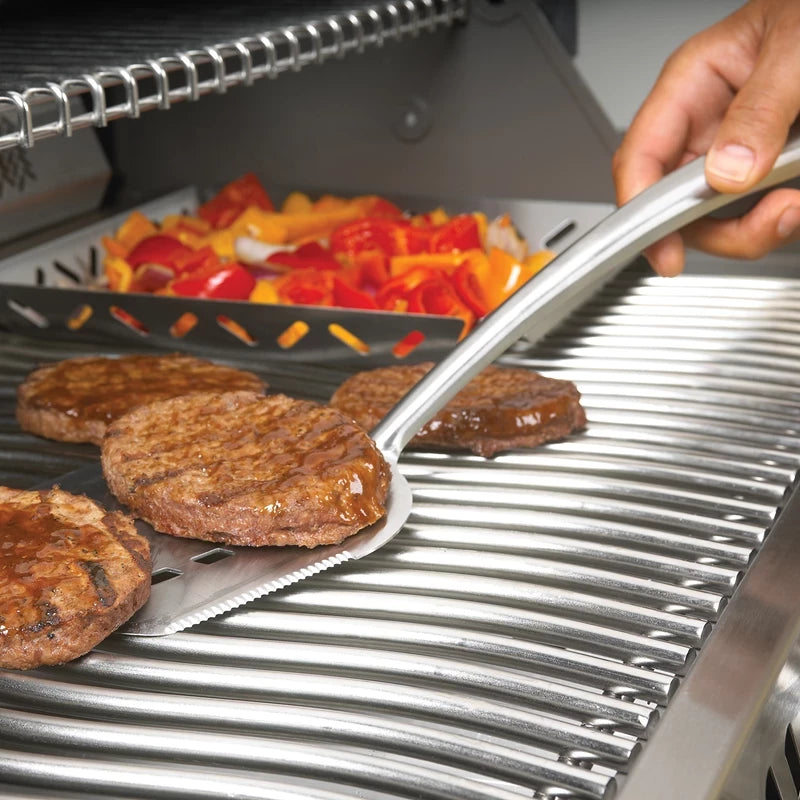 En hånd snur hamburgerbiffene på grillen med Napoleon PRO-grillspaden, mens skiver av rød og gul paprika steker i en form like ved. Dette uunnværlige grillutstyret gir perfekte grillresultater hver gang.