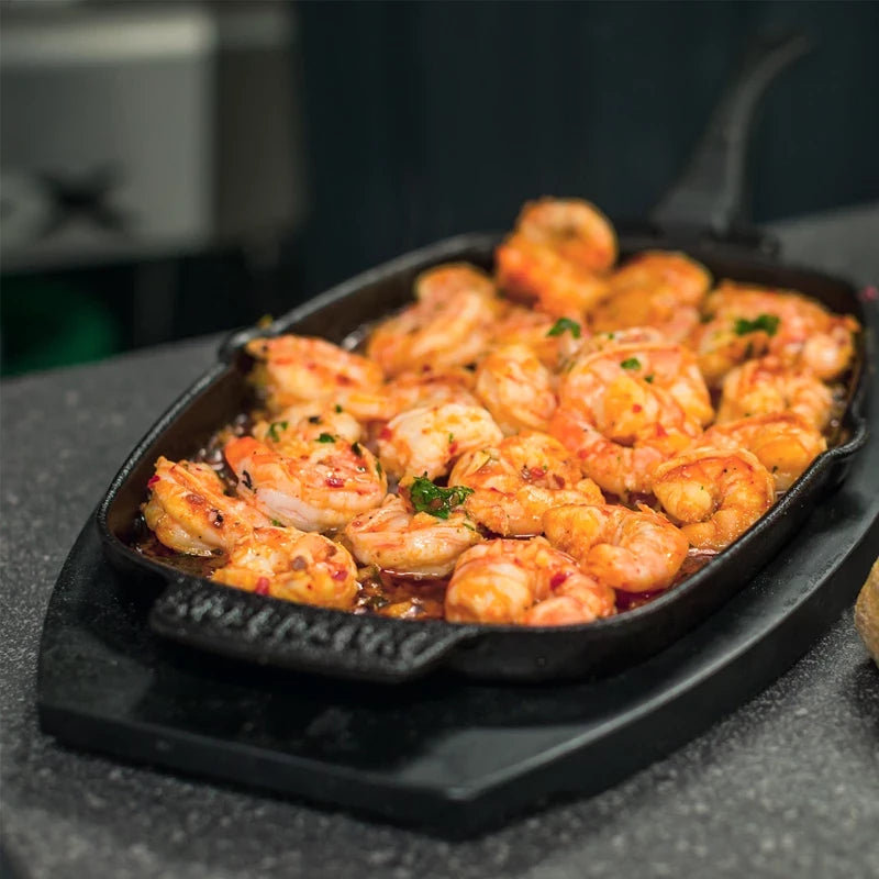 En Napoleon PRO Sizzle-panne i støpejern er fylt med kokte reker i saus og pyntet med hakkede urter. Den står på en mørk benkeplate.