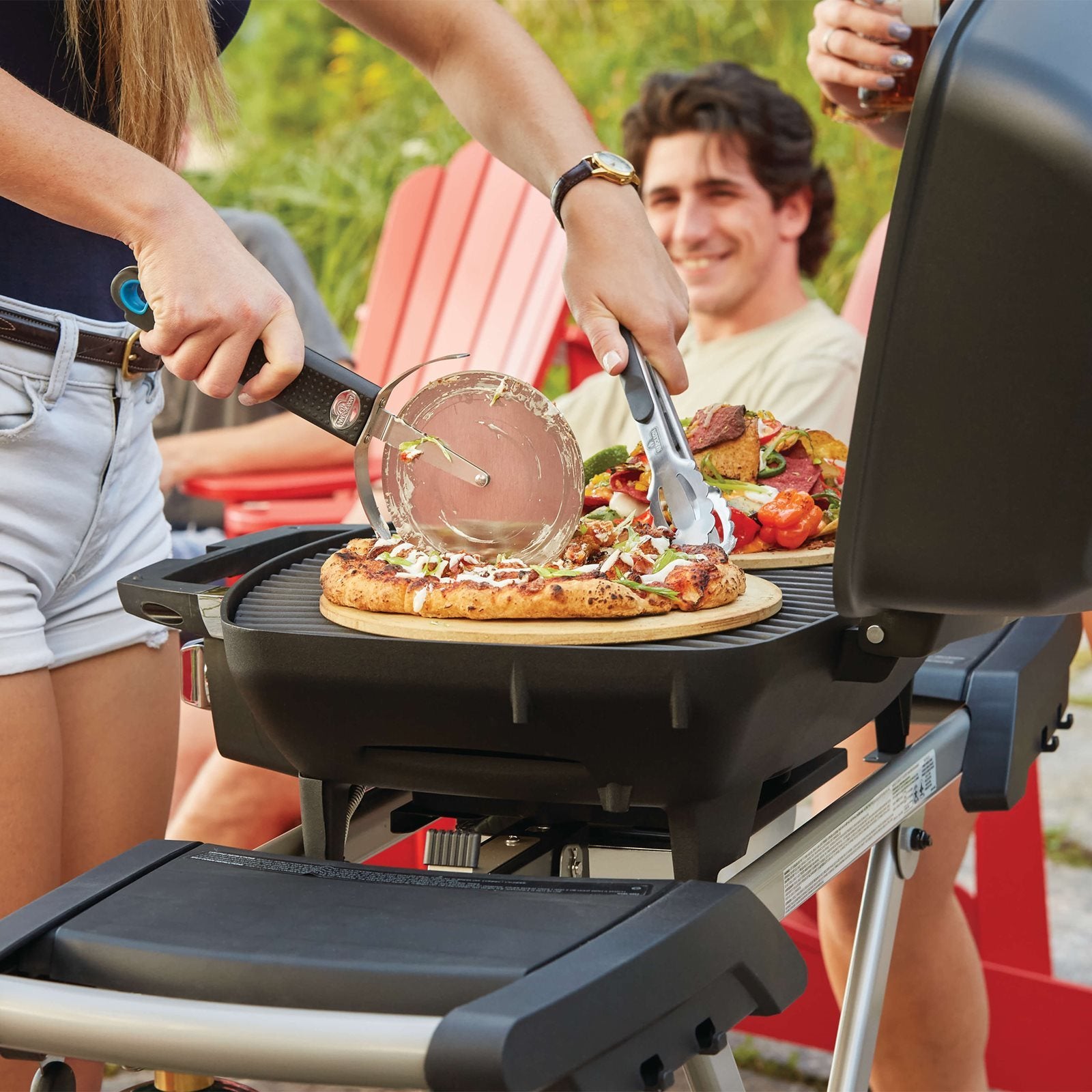 En person skjærer pizza på en Napoleon TravelQ PRO 285X Gassgrill mens en annen holder en tallerken med grillede grønnsaker; to personer sitter i røde Adirondack-stoler i nærheten.