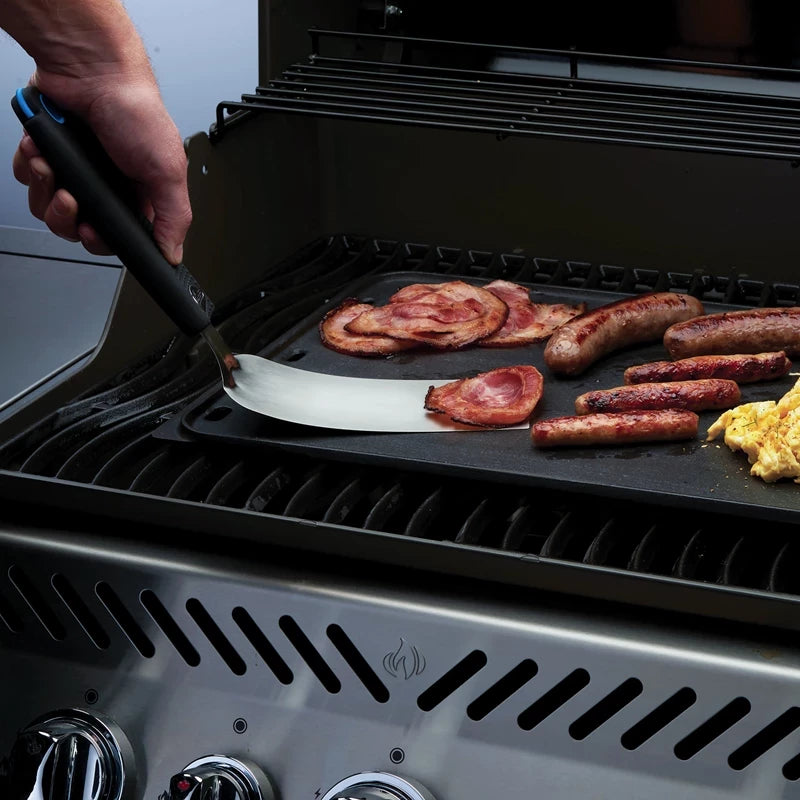 En person bruker en Napoleon Fleksibel Stekespade til å tilberede bacon, pølser og eggerøre på en stekeplategrill med flere kontrollknapper.