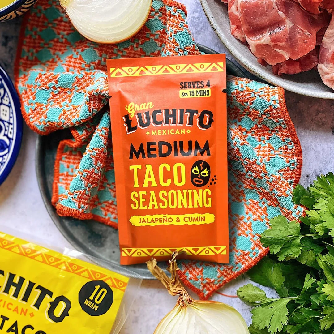 En 28 g-pakke med Gran Luchito Medium Tacokrydder ligger på et fargerikt klede ved siden av rått kjøtt, løk, koriander og taco-wraps - perfekt for å tilføre autentisk meksikansk smak til måltidet ditt.