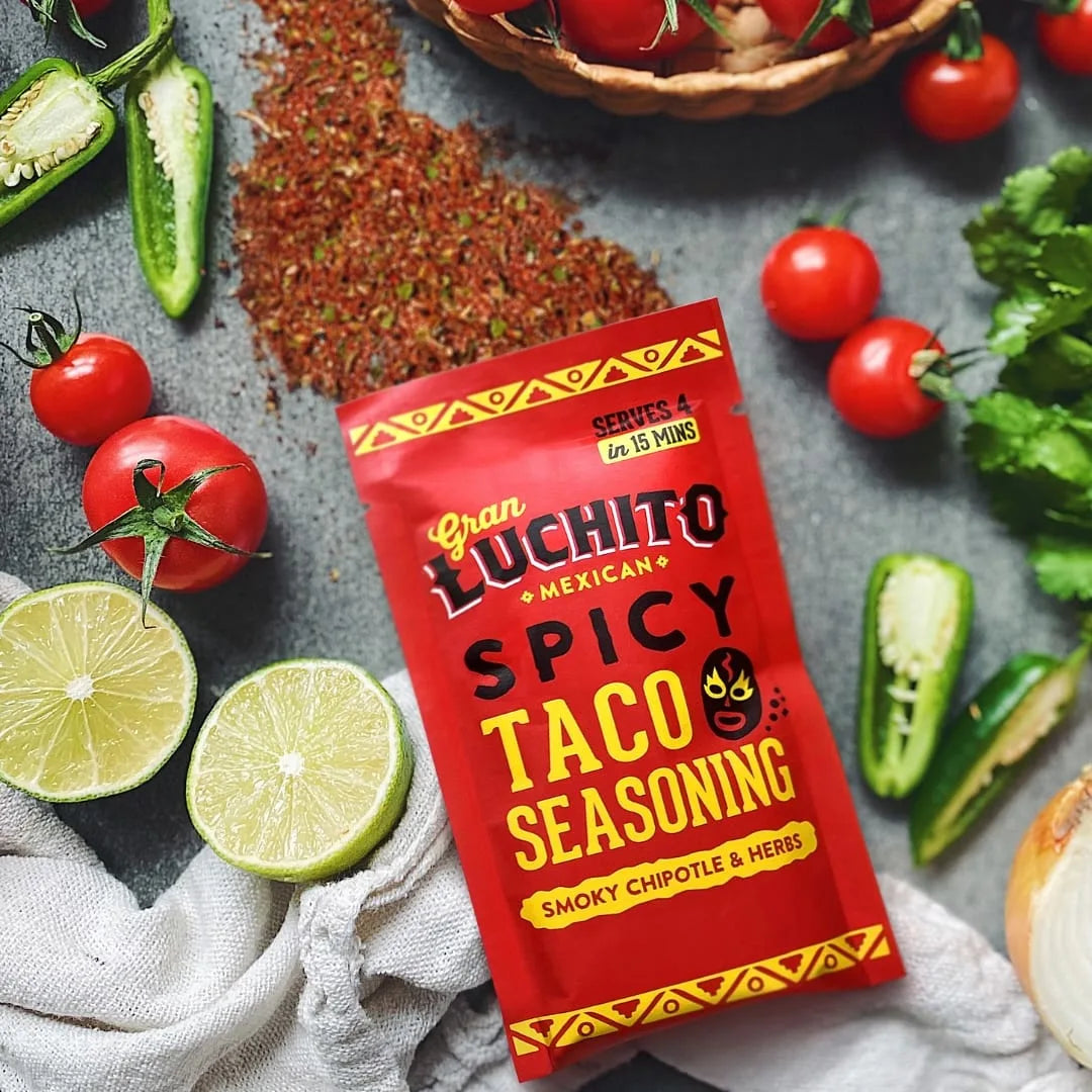 En pakke Gran Luchito Spicy Taco Seasoning 28 g er omgitt av friske limefrukter, tomater, jalapeños, koriander og en lys klut på en grå overflate, som fremhever denne smakfulle meksikanske krydderblandingen.