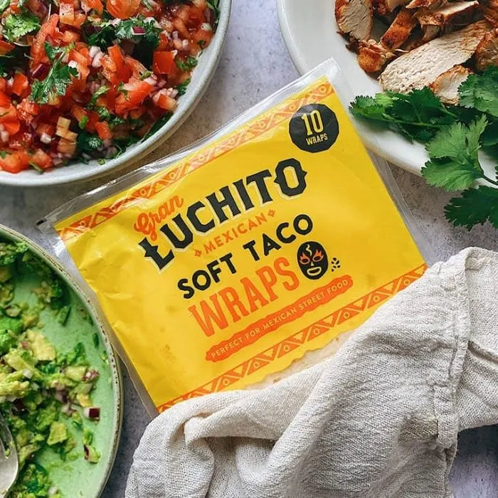 En pakke Gran Luchito Soft Taco Wraps på 300 g plassert ved siden av skåler med guacamole, pico de gallo, grillet kylling og fersk koriander – perfekt for å lage autentisk meksikansk gatemat hjemme.