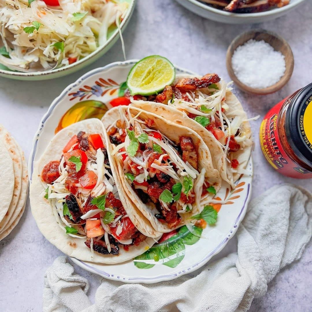 Tre tacoer laget med Gran Luchito Soft Taco Wraps 300 g er fylt med grillet kjøtt, revet ost, tomater, urter og salat, og serveres på en tallerken med en lime-skive. En bolle med salat, salt og en hermetikkboks står ved siden av for å gi den perfekte opplevelsen av meksikansk gatemat.