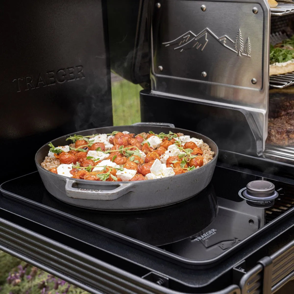 Kirsebærtomater, ost og friske urter syder i en rund stekepanne på Traeger Timberline XL pelletsgrill, og dampen stiger opp når grillens metallokk kommer til syne - et perfekt øyeblikk for utendørs matlaging.