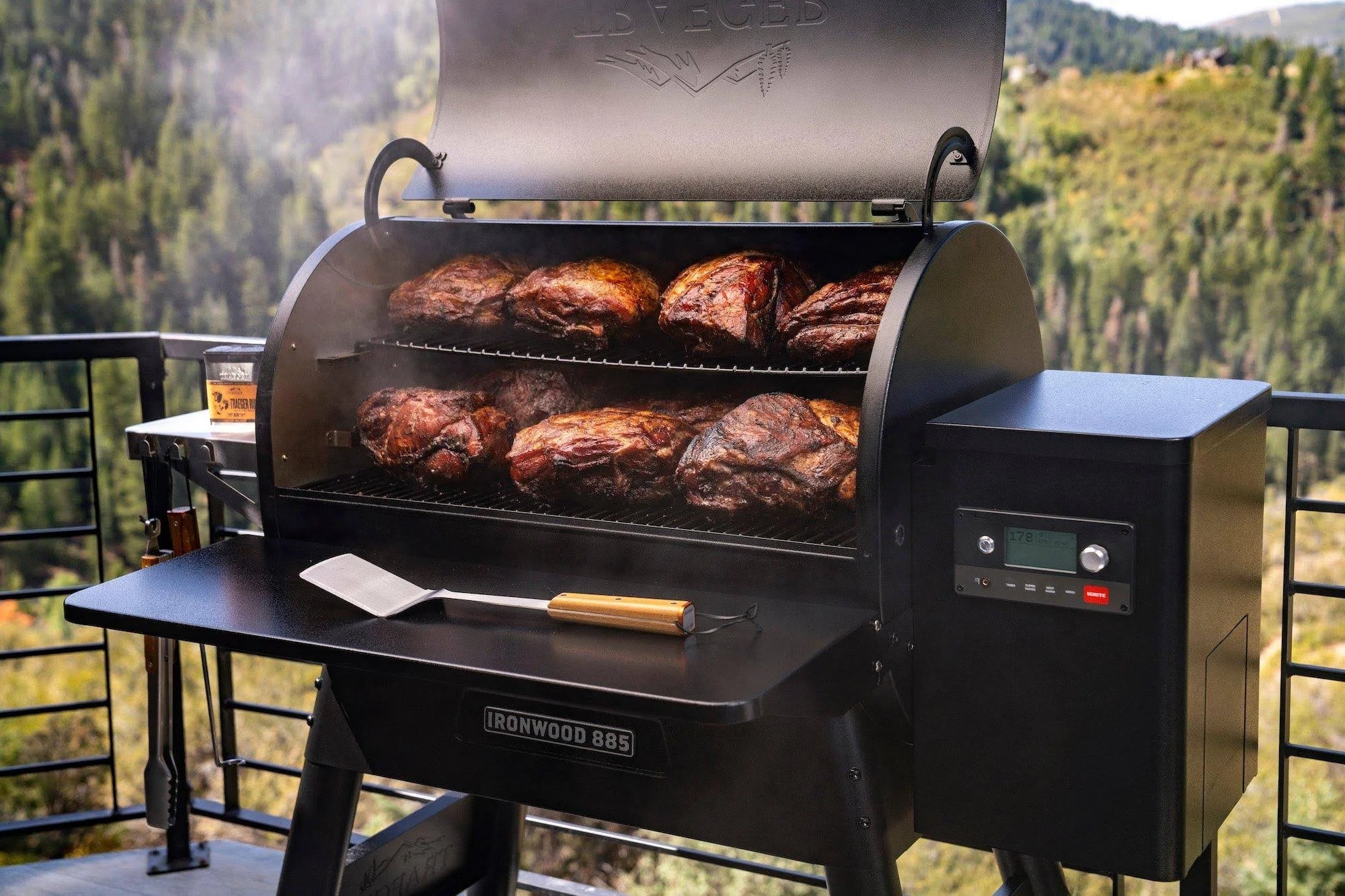 En Traeger Ironwood D2 885 pelletsgrill i svart, merket Ironwood 885, tilbereder store kjøttstykker på to rister. Den står på en terrasse med utsikt over skogkledde fjell. Inkluderer WiFIRE-teknologi.