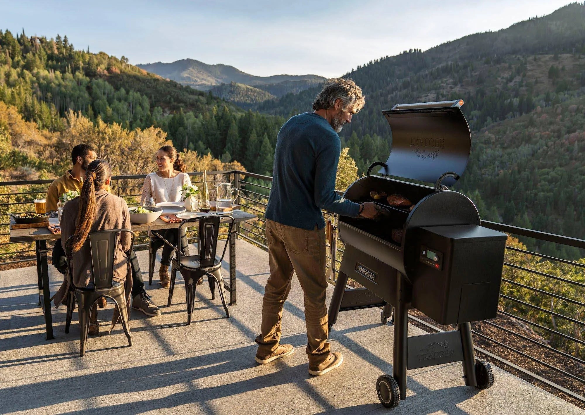 En mann griller mat på en Traeger Ironwood D2 885 mens tre personer sitter ved et bord på en uteplass med utsikt over skogkledde fjell.