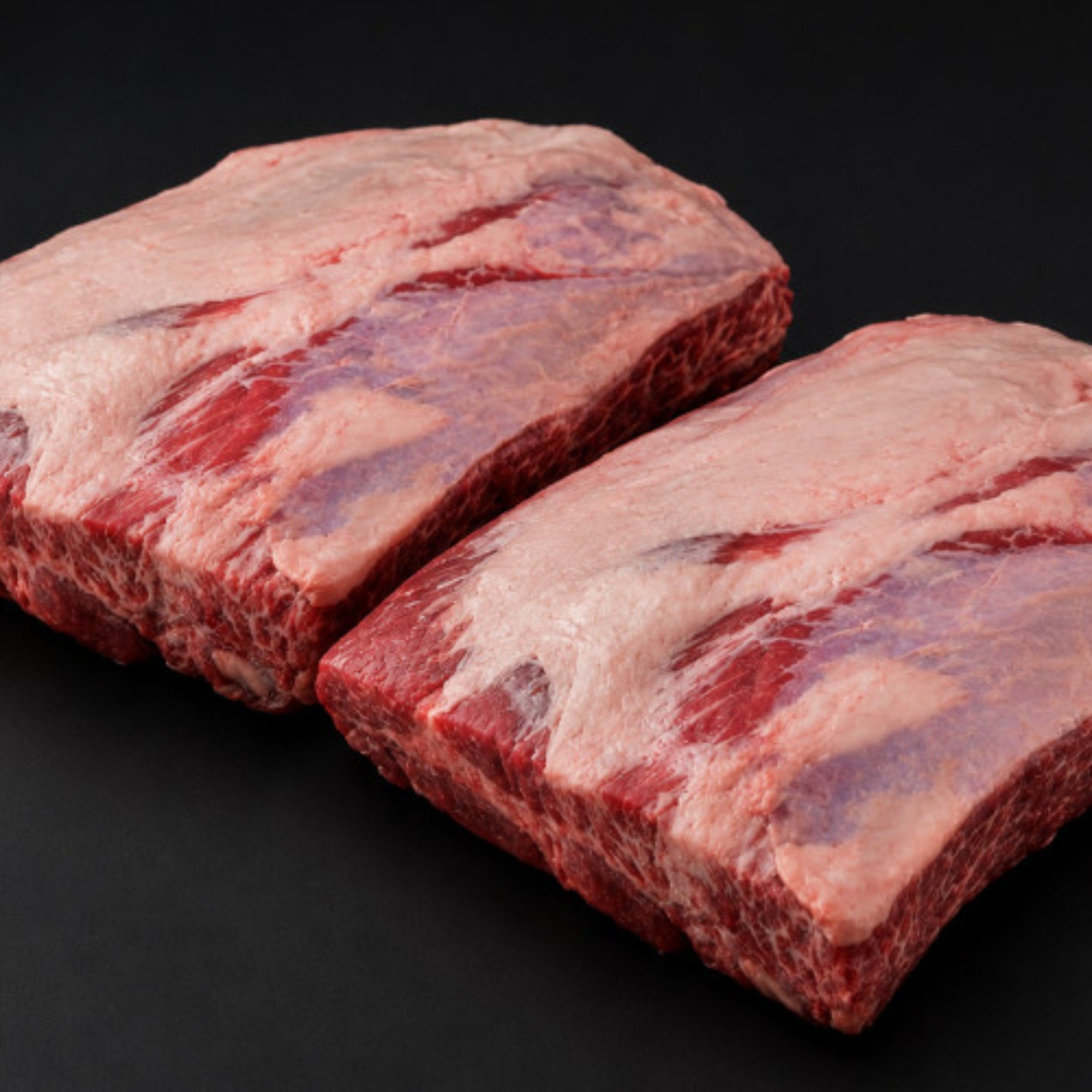 To rå Storfe Short Ribs Angus fra Irland, priset til kr 250 per kg, med synlig marmorering og fettkappe er plassert side om side på en svart flate - perfekt til grillmat.