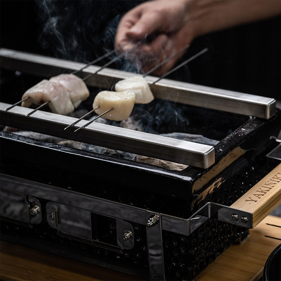 En hånd holder en YAKINIKU BBQ Spyd i Rustfritt Stål 1,0 mm 20stk over en rektangulær bordgrill, med røyk som stiger opp fra den sydende maten.