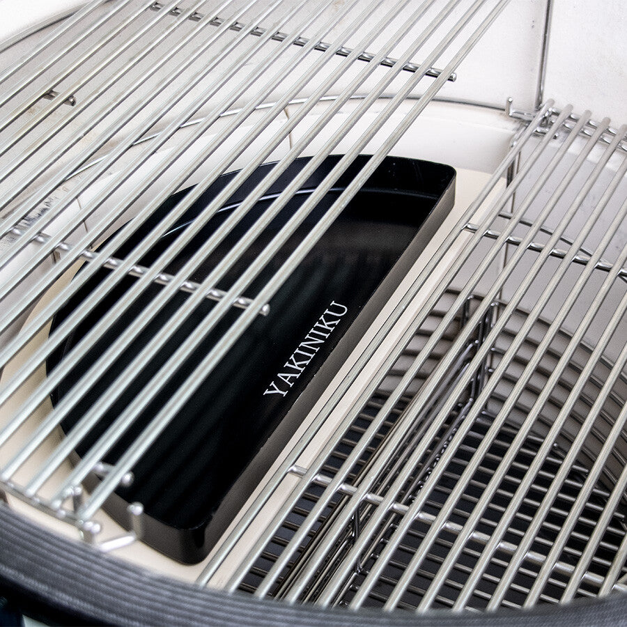 Nærbilde av tomme grillstativer i metall inne i en kamadogrill, med en YAKINIKU Halvmåne Dryppfat XLARGE / LARGE for indirekte grilling plassert under stativene.