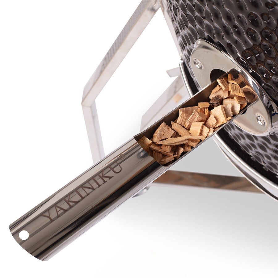 Nærbilde av en metallskje merket YAKINIKU fylt med flis som settes inn i en keramisk grill, og som viser frem proffsettet som følger med YAKINIKU Kamado XXLARGE Complete.