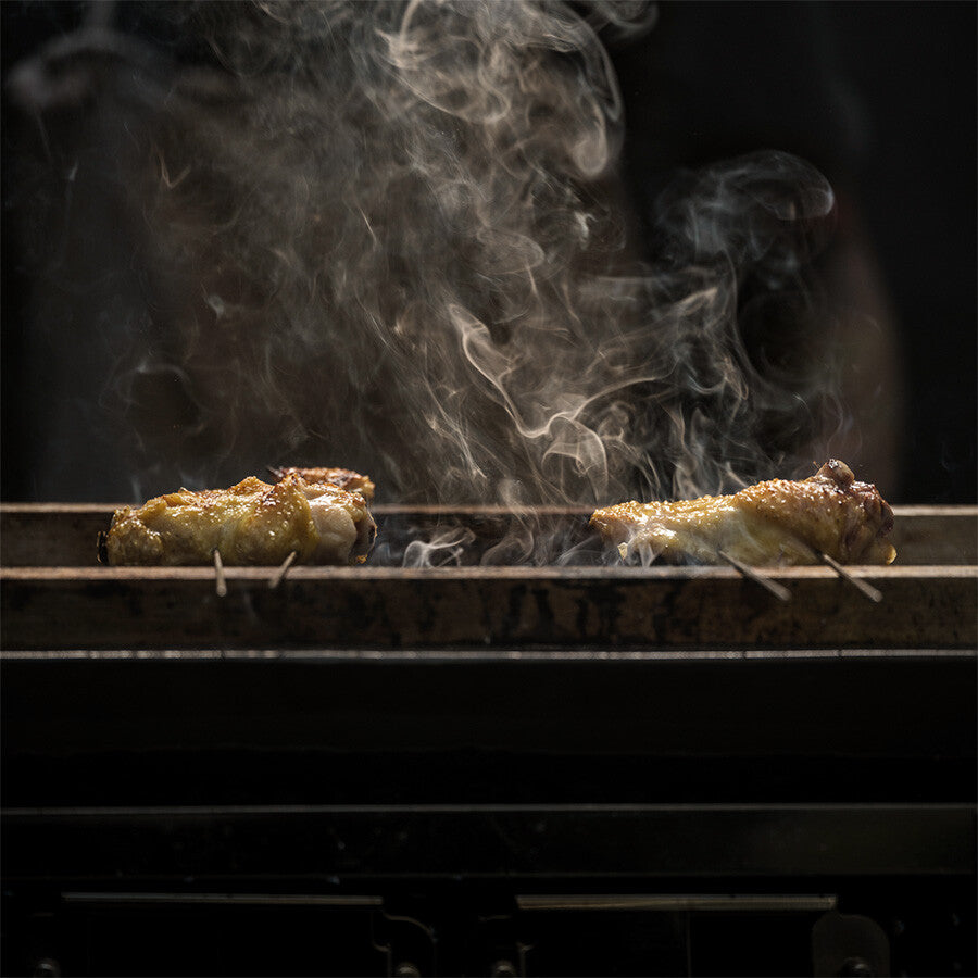To kyllingstykker grilles på YAKINIKU LARGE Rectangular Shichirin | Konro & Yakitori Grill, med røyk som stiger opp mot en mørk bakgrunn.