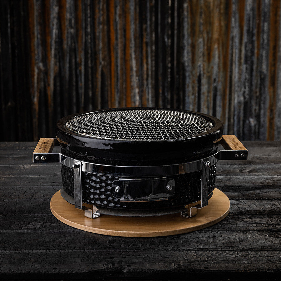 YAKINIKU Round Shichirin Japansk Bordgrill har et rundt, svart design med metallhåndtak, nettingrist og står på en trebase med en rustikk bakgrunn.