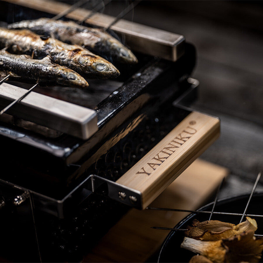 Tre hele fisk grilles på metallspyd over en YAKINIKU Rectangular Shichirin | Konro & Yakitori Grill, med "YAKINIKU"-logoen godt synlig på trehåndtaket.