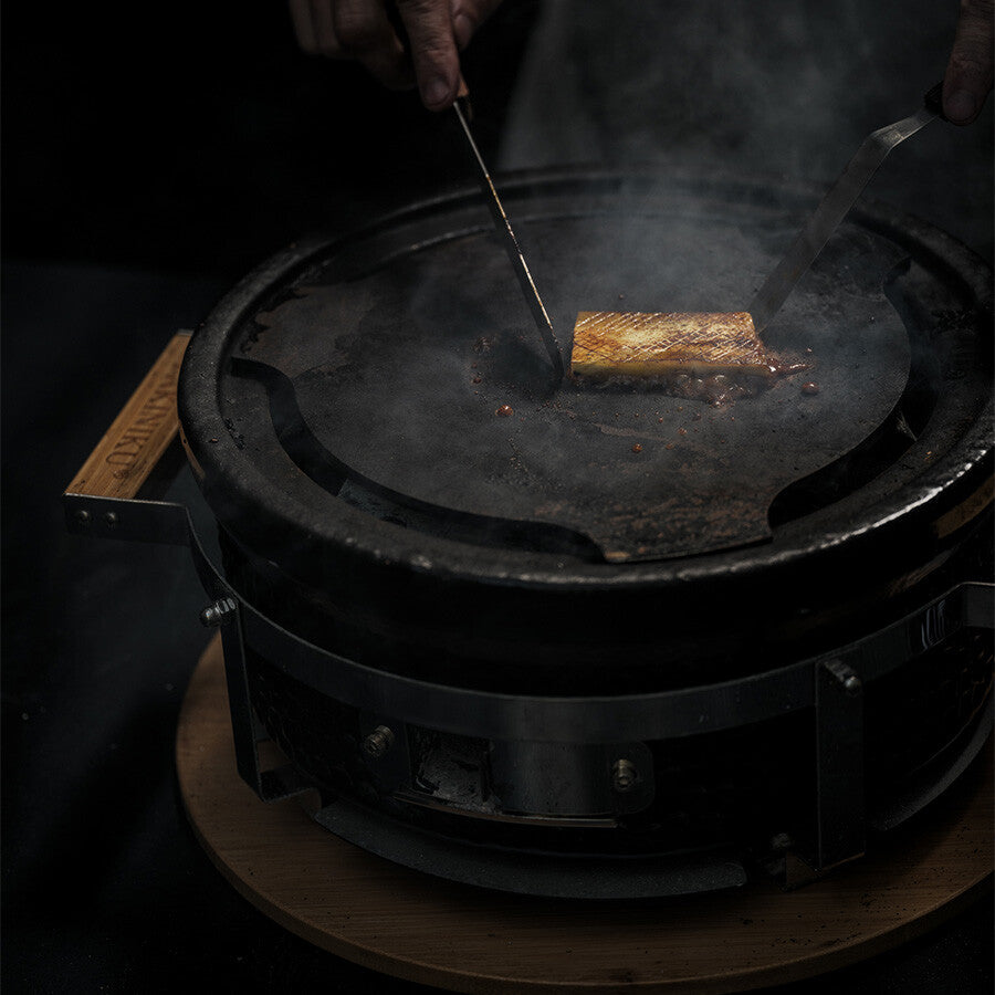 Noen bruker redskaper til å tilberede et rektangulært stykke mat på en rund, svart YAKINIKU Shichirin Teppanyaki Plate Rund (Medium) grill, mens røyken stiger opp.