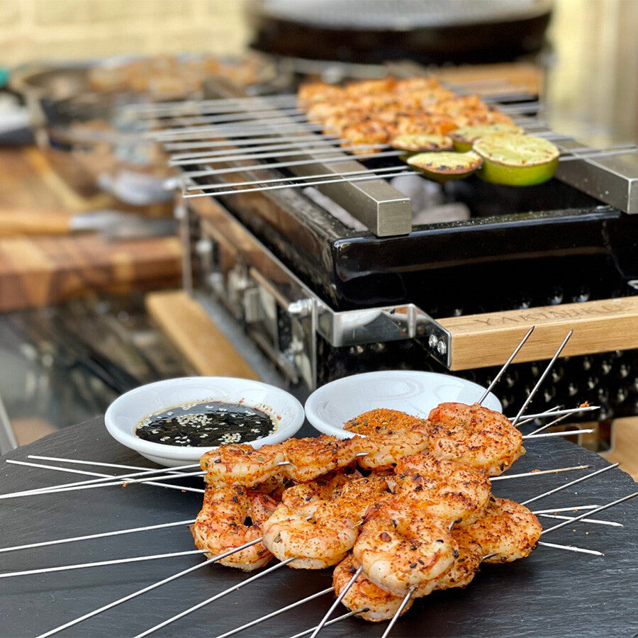 Krydrede grillede rekespyd serveres på en svart tallerken med to sauser, mens flere spyd og grønnsaker tilberedes på en YAKINIKU Shichirin Yakitori Bars | MEDIUM, som fremkaller en autentisk japansk grillopplevelse.