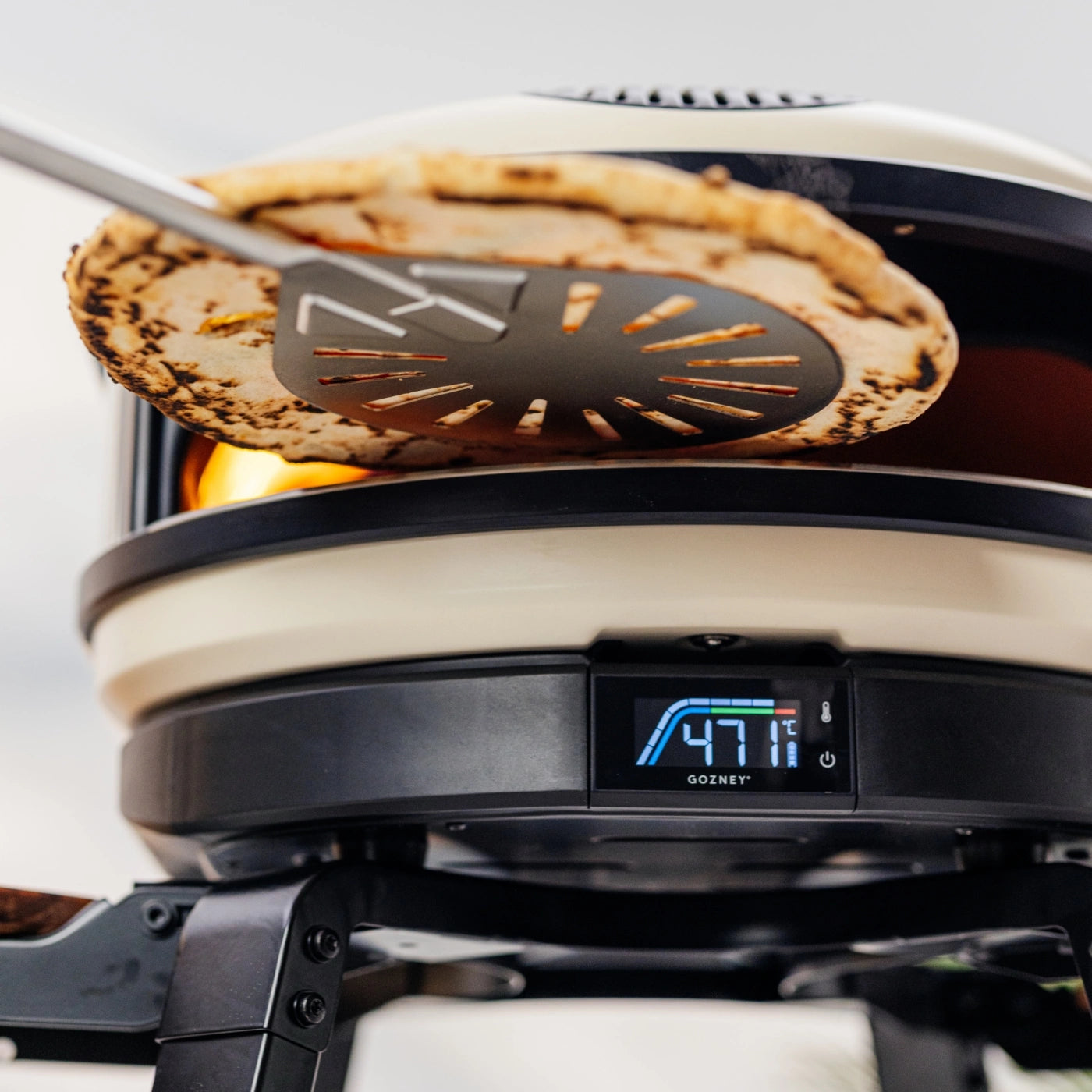 En pizza legges inn i en Gozney-pizzaovn, der displayet viser 477 °C, ved hjelp av den rustfrie stålspaden «Gozney Balance Turning Peel».