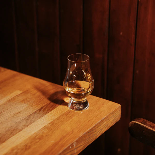 Et Glencairn Whisky Glass 190 ml fra Stölzle Lausitz 6-delt sett står på kanten av et trebord, med en mørk trevegg i bakgrunnen.