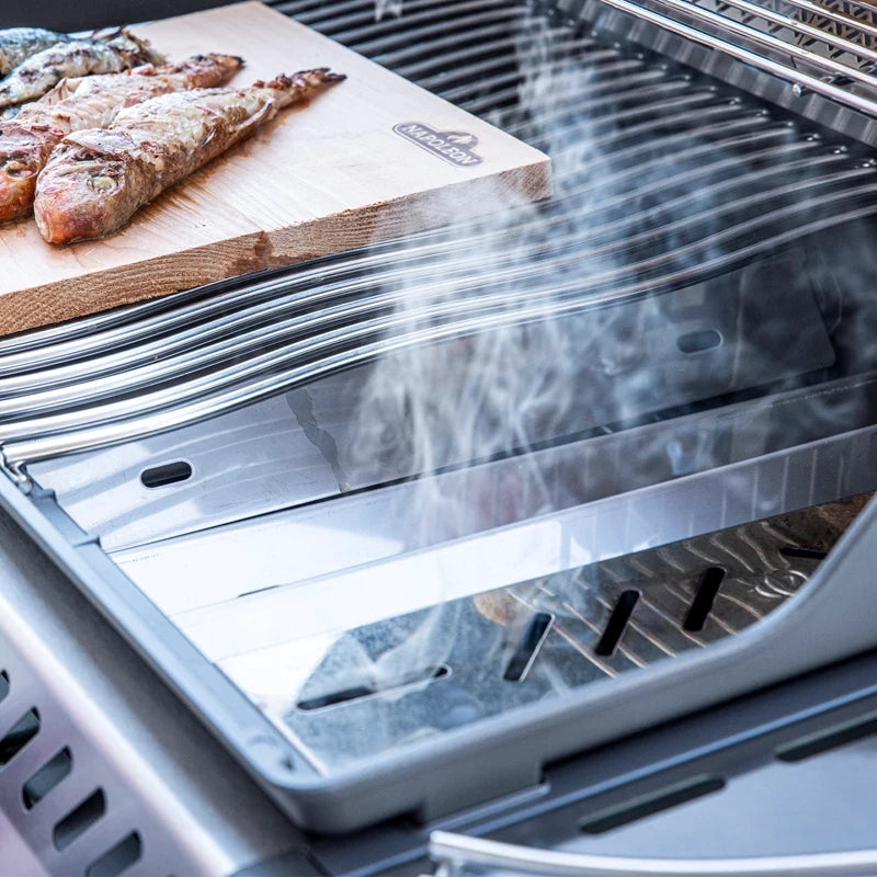 En Napoleon Sear Plates Smoker Box - røykeeske på en metallgrill avgir rik røyksmak gassgrillsmak, med røyk som stiger opp mens den kokte fisken hviler på et skjærebrett av tre i nærheten.