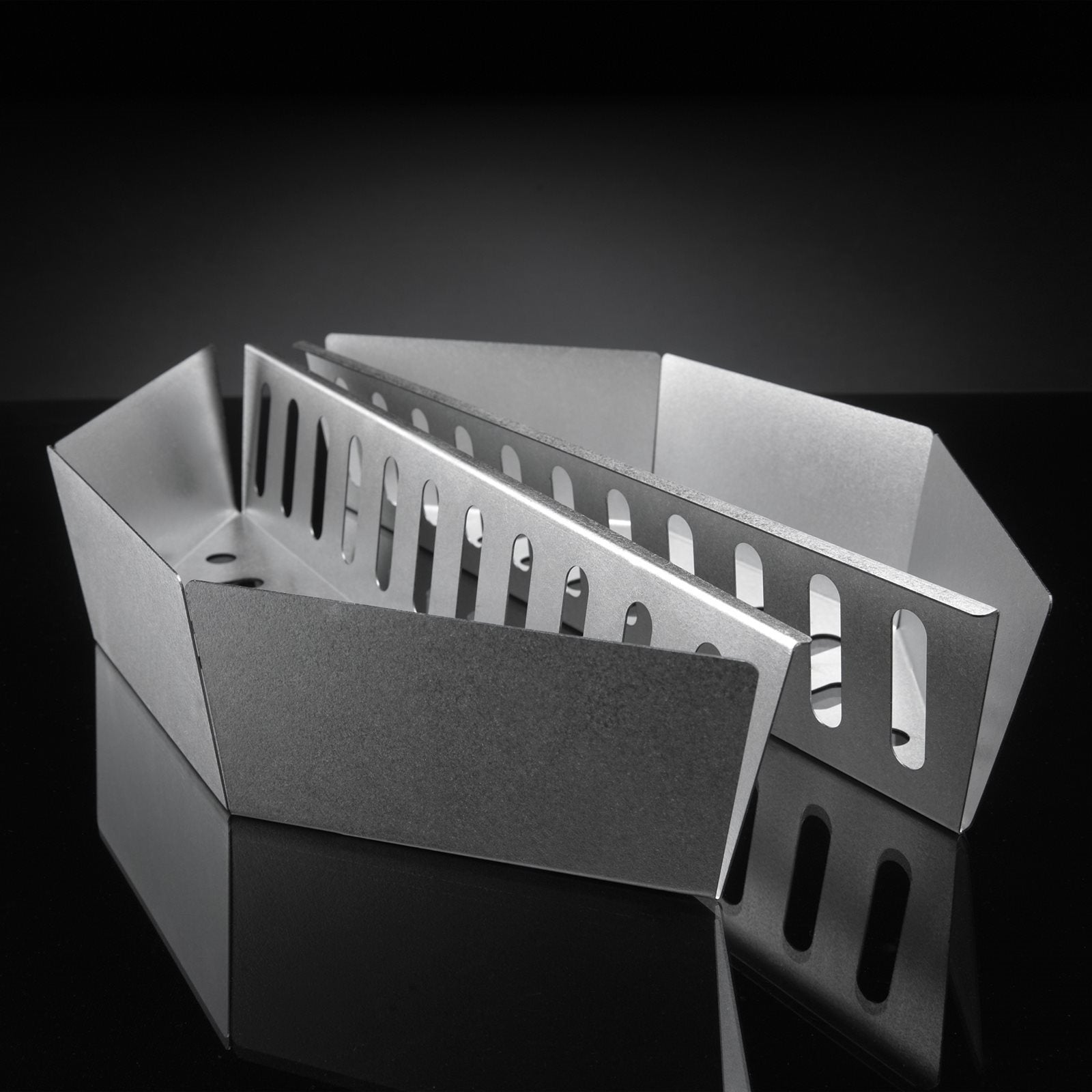 To Napoleon kurver til grillkull for kulegrill, 2 stk. i sølv - en hel og en med rektangulære utskjæringer - er plassert på en reflekterende svart flate mot en mørk bakgrunn.